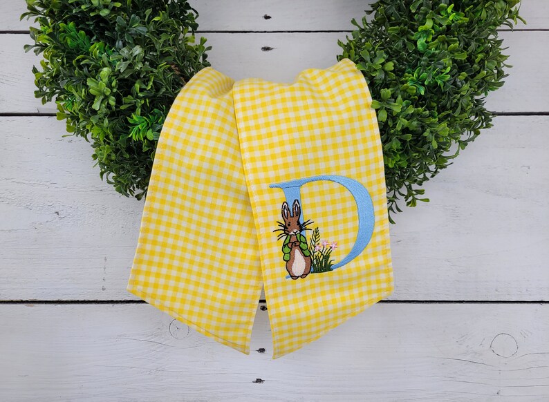 May include: Yellow and white gingham fabric with a blue embroidered letter "D" and a bunny wearing a green jacket. The bunny is standing in a patch of flowers. The fabric is draped over a green wreath.