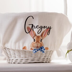 May include: White wicker Easter basket with a white linen cover featuring a brown bunny with a blue bow tie and the name "Gregory" printed in black.