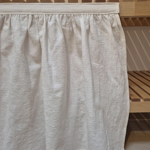 May include: White cotton valance with a ruffled edge, hanging on a wooden shelf.