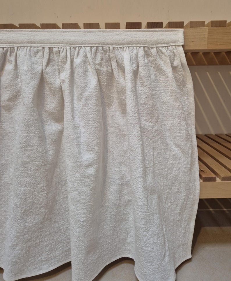 May include: White cotton valance with a ruffled edge, hanging on a wooden shelf.