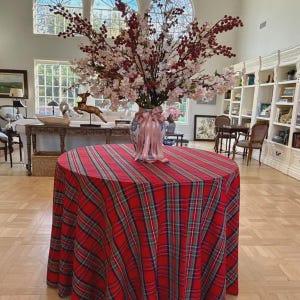 May include: A round table covered with a red, green, and blue plaid tablecloth. Centered on the table is a ceramic vase filled with tall, branching stems featuring small white blossoms and clusters of dark red berries, tied with a pink ribbon.