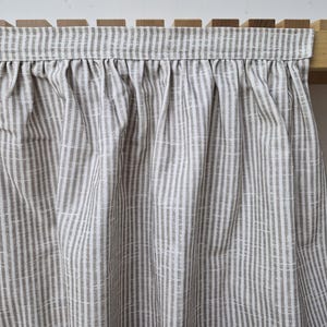 May include: A gathered, light gray and white striped curtain panel. The fabric has a vertical stripe pattern with a slightly textured appearance. The curtain is attached to a wooden shelf unit, suggesting a home decor item.