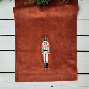 May include: A rust-colored velvet table runner with an embroidered nutcracker design. The nutcracker is wearing a blue and white uniform with a gold crown.
