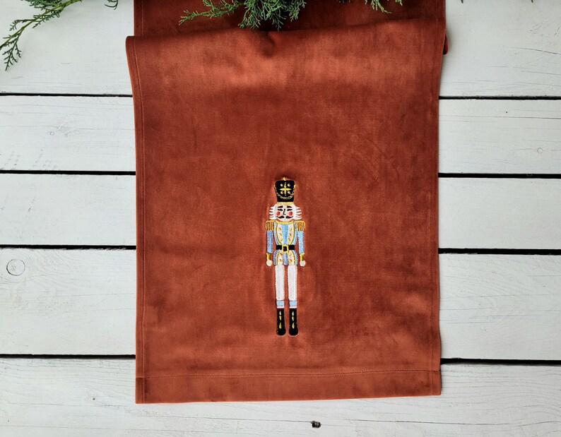 Op de afbeelding: Een roestkleurige fluwelen tafelkleed met een geborduurd notenkrakerontwerp. De notenkraker draagt een blauw-wit uniform met een gouden kroon.