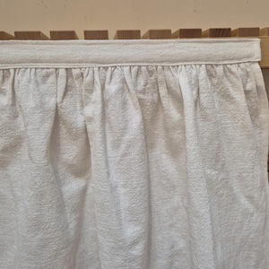 May include: White cotton curtain with a ruffled hem, hanging from a wooden shelf.
