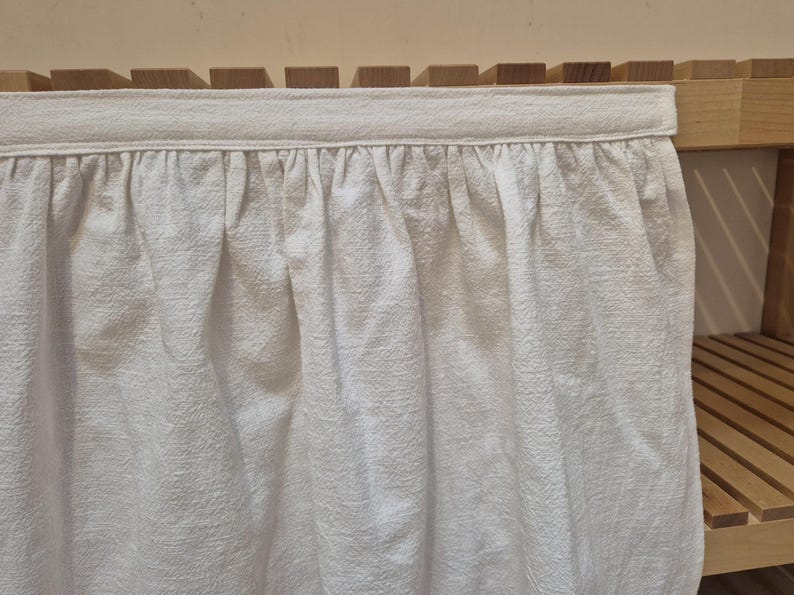 May include: White cotton curtain with a ruffled hem, hanging from a wooden shelf.