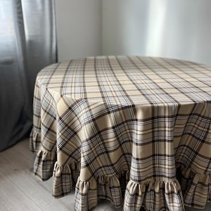May include: A round tablecloth featuring a beige, brown, and light blue plaid pattern. The fabric drapes over a circular table and includes a gathered ruffle trim along the bottom edge, creating a layered effect around the perimeter.