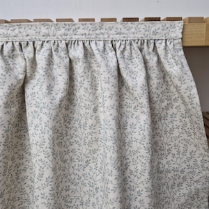 May include: A pale fabric curtain featuring a delicate pattern of grey leaves and branches. The curtain is gathered at the top, creating soft folds. The fabric is draped over a wooden structure.