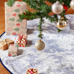 May include: A decorated Christmas tree with ornaments and wrapped gifts. The tree skirt is white with a blue toile pattern and a dark blue border. Some gifts are wrapped in brown paper with Christmas tree and heart patterns.