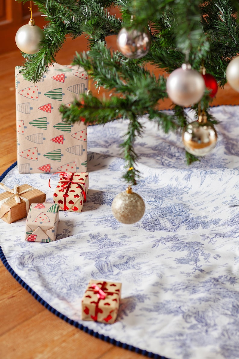 May include: A decorated Christmas tree with ornaments and wrapped gifts. The tree skirt is white with a blue toile pattern and a dark blue border. Some gifts are wrapped in brown paper with Christmas tree and heart patterns.