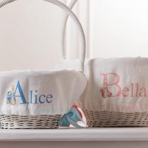 May include: Two white wicker Easter baskets with personalized fabric liners. The basket on the left has a blue bunny and the name "Alice" embroidered on it. The basket on the right has a pink bunny and the name "Bella" embroidered on it.