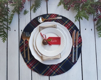 May include: A festive table setting with a plaid placemat, white plates, and silverware. A red tag with the name "Kate" is attached to a small bell. Greenery and berries decorate the background.