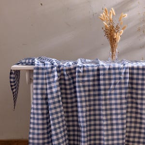 May include: A white table covered with a blue and white gingham tablecloth. A clear glass vase with dried flowers is on the table. The tablecloth drapes over the edge. The background is a neutral-toned wall. The table is set on a wooden floor.
