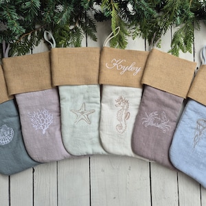 May include: Six personalised Christmas stockings with a burlap cuff and a linen body. Each stocking features a different embroidered sea creature, including a starfish, a seahorse, a crab, a jellyfish, a seashell, and coral.
