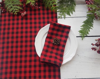 May include: A red and black buffalo check table runner and matching napkin are displayed on a white wooden surface. A white plate holds the folded napkin, adorned with a small pinecone. Greenery and red berries add a festive touch.