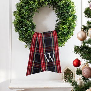 May include: A green boxwood wreath with a red and black plaid ribbon tied around it. The ribbon has a white letter "W" embroidered on it. The wreath is hanging on a white wall above a white shelf with a small gold house figurine on it. A Christmas tree with ornaments is in the background.