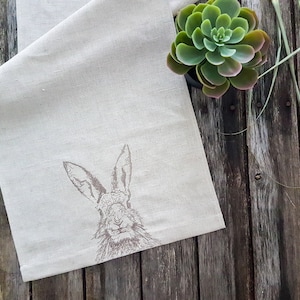 Può includere: Un runner da tavolo in lino bianco con un'immagine ricamata marrone di una testa di coniglio.