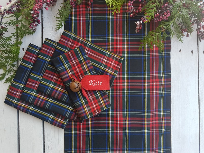 May include: A set of four red, black, green, and yellow plaid napkins with a matching plaid table runner. The napkins are folded and stacked on top of each other. The table runner is laid out flat. A small red tag with the name "Kate" is attached to the top napkin.