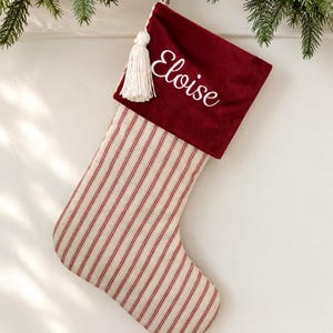 Puede incluir: Un calcetín navideño con cuerpo de tela a rayas color crema y rojo y un puño de terciopelo rojo oscuro. El puño está bordado con el nombre Eloise en letra blanca y tiene una borla blanca decorativa colgando de la esquina superior.