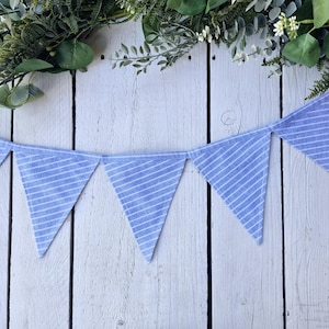 Puede incluir: Una guirnalda de banderines a rayas azules y blancas. Las banderas triangulares están unidas y cuelgan sobre un fondo de madera blanca. La vegetación y las flores blancas están en la parte superior de la imagen.