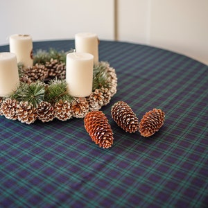 May include: A Christmas wreath with four white candles, pinecones, and faux evergreen branches. The wreath sits on a round table covered with a blue and green plaid tablecloth. Three brown pinecones are scattered on the table.