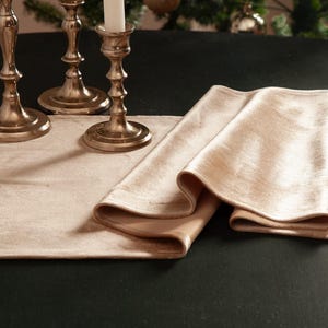 May include: A folded champagne-colored velvet table runner rests on a dark surface, with three silver candle holders and a Christmas tree in the background. The runner's texture is smooth and reflective.