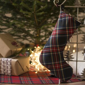 Op de afbeelding: Een rode en zwarte geruite kerstsok die aan een metalen standaard hangt met een wit, pluche oppervlak op de achtergrond. De sok is versierd met een wit en geel geruit patroon.