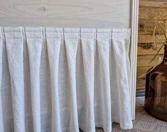 May include: A white linen bed skirt with a pleated design. The fabric hangs in vertical folds, creating a textured appearance. The bed skirt is set against a light-colored wooden headboard and a wooden wall.