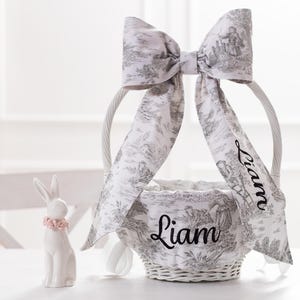 May include: White wicker Easter basket with a gray and white toile ribbon bow. The ribbon is tied in a bow and the ends are embroidered with the name "Liam".