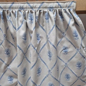 May include: A cream-coloured fabric with a repeating blue floral and diamond pattern. The fabric is gathered at the top, creating soft folds. The design features stylized flowers and a lattice-like structure.
