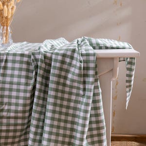 Puede incluir: Una mesa de madera blanca cubierta con un mantel a cuadros verdes y blancos. Un jarrón con flores secas está sobre la mesa. El mantel cuelga del borde de la mesa.