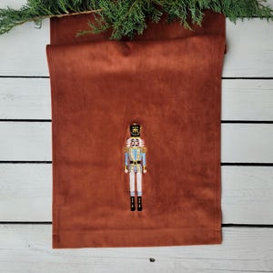 Op de afbeelding: Een bruine fluwelen tafelrunner met een geborduurd notenkrakerontwerp. De notenkraker draagt een blauw en wit uniform met een gouden kroon.