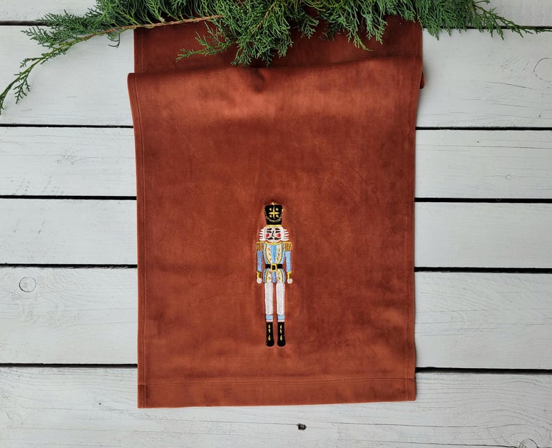 Op de afbeelding: Een bruine fluwelen tafelrunner met een geborduurd notenkrakerontwerp. De notenkraker draagt een blauw en wit uniform met een gouden kroon.