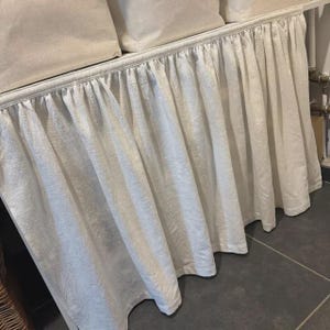 Può includere: Una gonna da letto color crema con un design a balze e arricciature. Il tessuto sembra essere lino o un materiale strutturato simile. La gonna è attaccata a un telaio del letto con cuscini color crema abbinati visibili.