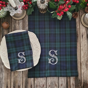 Peut inclure: Un set de table et de serviette en tissu écossais vert et bleu avec une lettre "S" brodée en blanc sur chaque pièce. Le set est posé sur une table en bois avec des baies rouges et de la verdure.