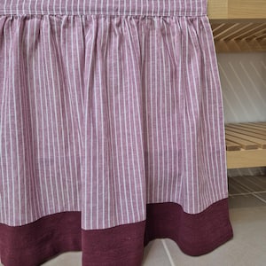 May include: A burgundy and white striped curtain with a solid burgundy border at the bottom. The curtain is hanging in front of a wooden shelf. The fabric has vertical white stripes on a burgundy background.