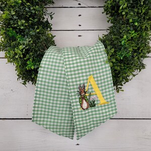 May include: A green and white gingham ribbon with an embroidered bunny and the letter "A" in yellow. The ribbon is draped over a green wreath, creating a festive Easter decoration.
