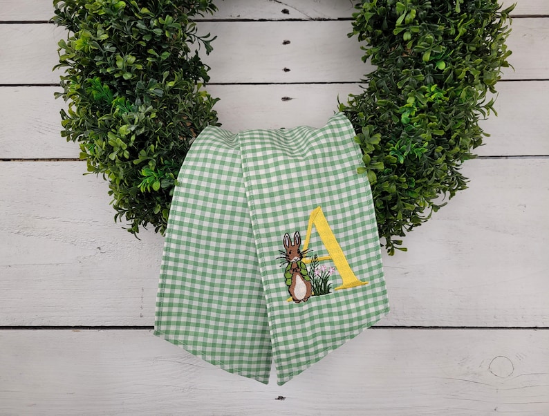 May include: A green and white gingham ribbon with an embroidered bunny and the letter "A" in yellow. The ribbon is draped over a green wreath, creating a festive Easter decoration.