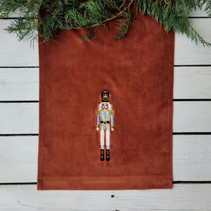 Op de afbeelding: Een roestkleurige tafelrunner met een geborduurd notenkrakerontwerp. De notenkraker draagt een blauw en gouden uniform met een zwarte hoed.