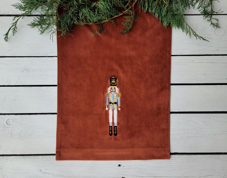 Op de afbeelding: Een roestkleurige tafelrunner met een geborduurd notenkrakerontwerp. De notenkraker draagt een blauw en gouden uniform met een zwarte hoed.