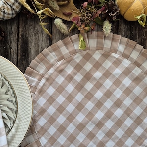 Puede incluir: Un mantel individual redondo de tejido de cuadros beige y blanco con un borde rizado. El mantel individual está hecho de tela y tiene un estilo informal de granja.