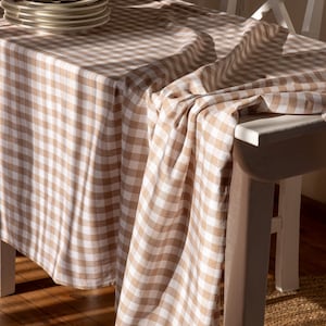 Puede incluir: Un mantel a cuadros beige y blanco sobre una mesa blanca. Una pila de platos, servilletas y un servilletero están sobre la mesa. El mantel es de tela suave y es adecuado para un comedor.
