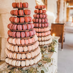 May include: A tiered stand with two levels of pink and white macarons, decorated with greenery. The stand is on a white dresser with a lace overlay.