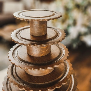 Può includere: Un porta torte in legno a più livelli con bordi a festoni. Il porta torte è in legno marrone scuro e ha un aspetto rustico.