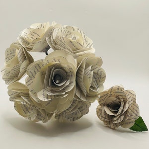 May include: A bouquet of five cream-colored paper roses made from book pages. The roses are arranged in a cluster, with one smaller rose with a green leaf stem to the side.