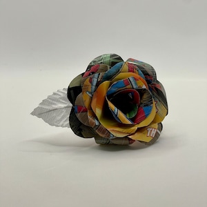 May include: A colorful paper rose made from comic book pages. The rose is yellow, red, blue, and green. It has a silver leaf.