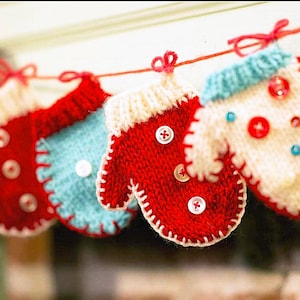 May include: A garland of knitted mittens in red, white and blue with white buttons. The mittens are strung on a red string and hang from a white surface.