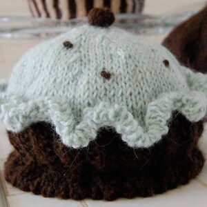 May include: A knitted cupcake hat with a light blue frosting and brown chocolate base. The hat has a ruffled edge and is decorated with brown sprinkles.