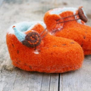 May include: A pair of orange knitted baby booties with blue and brown felt accents. The booties have a simple design and are perfect for a newborn.
