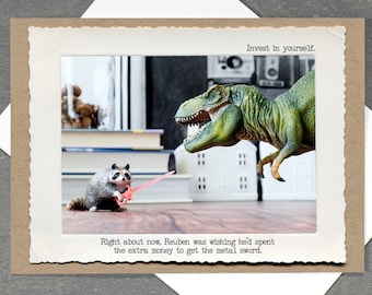 May include: Greeting card with a humorous scene. A green Tyrannosaurus Rex faces a raccoon holding a red lightsaber. Text reads "Invest in yourself" and "Right about now, Reuben was wishing he'd spent the extra money to get the metal sword."